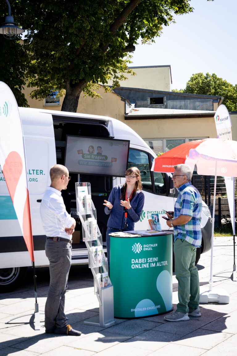 Ein Infostand des Projekts "Digitaler Engel" im Freien. Eine Mitarbeiterin mit Namensschild spricht mit zwei Männern vor einem weißen Infobus, der mit dem Logo des Projekts versehen ist. Auf einem Bildschirm am Bus ist der Schriftzug "Sie fragen. Wir antworten!" zu sehen. Neben dem Stand steht ein Prospektständer mit Flyern, sowie ein Sonnenschirm mit dem Logo des Digitalen Engels. Die Szene spielt sich auf einem sonnigen Platz unter einem Baum ab.