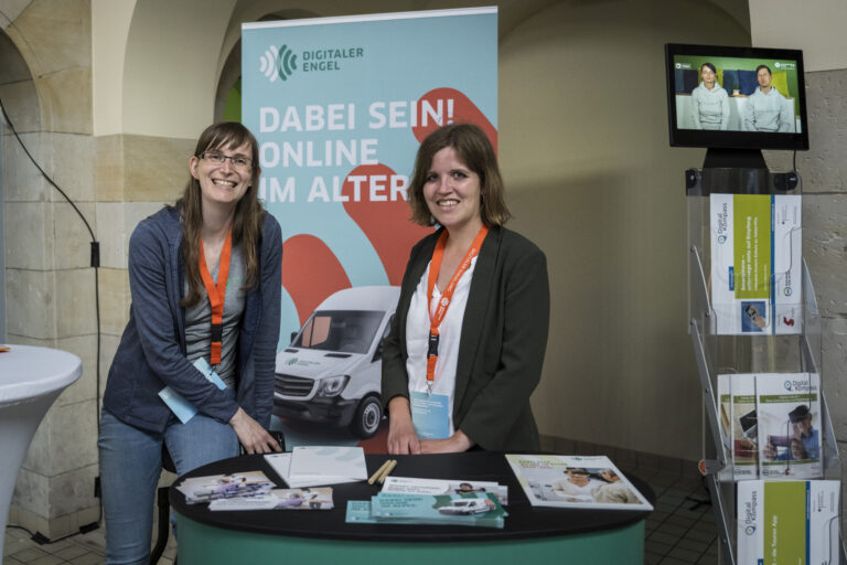 Zwei Frauen stehen an einem Infostand des Projekts 'Digitaler Engel'. Die Frau links trägt eine Brille, ein graues Oberteil und eine dunkelblaue Jacke. Die Frau rechts trägt einen dunkelgrünen Blazer über einem weißen Oberteil. Beide tragen orangefarbene Lanyards mit Namensschildern. Auf dem Tisch vor ihnen liegen Flyer und Broschüren. Im Hintergrund ist ein großes Banner mit der Aufschrift 'DABEI SEIN! ONLINE IM ALTER' sowie das Logo des Projekts zu sehen. Rechts neben dem Stand steht ein Prospektständer mit weiteren Informationsmaterialien, und darüber befindet sich ein Bildschirm mit einem Video.